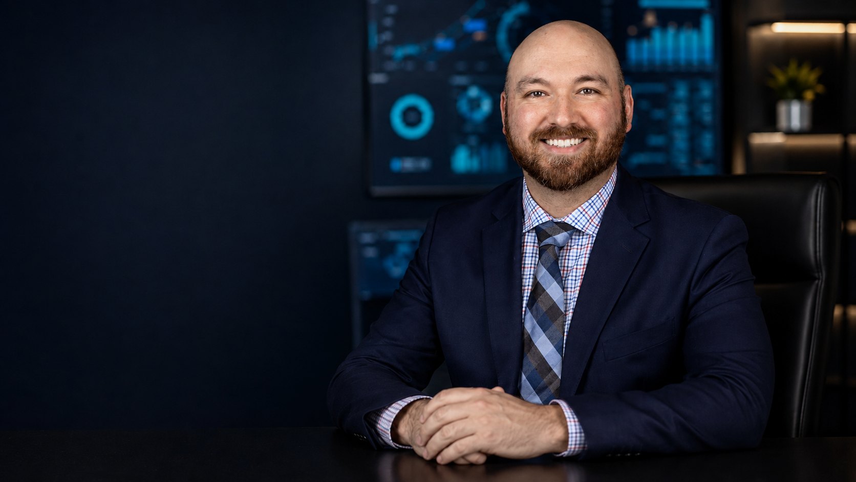 Christopher Cook in a navy suit and checked shirt, seated at a desk in front of data-visualization screens